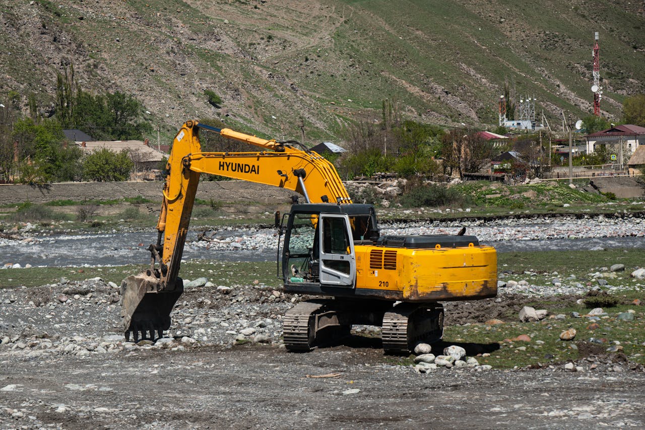 Yellow Hyundai excavator working on a rocky terrain near a river in a rural area.